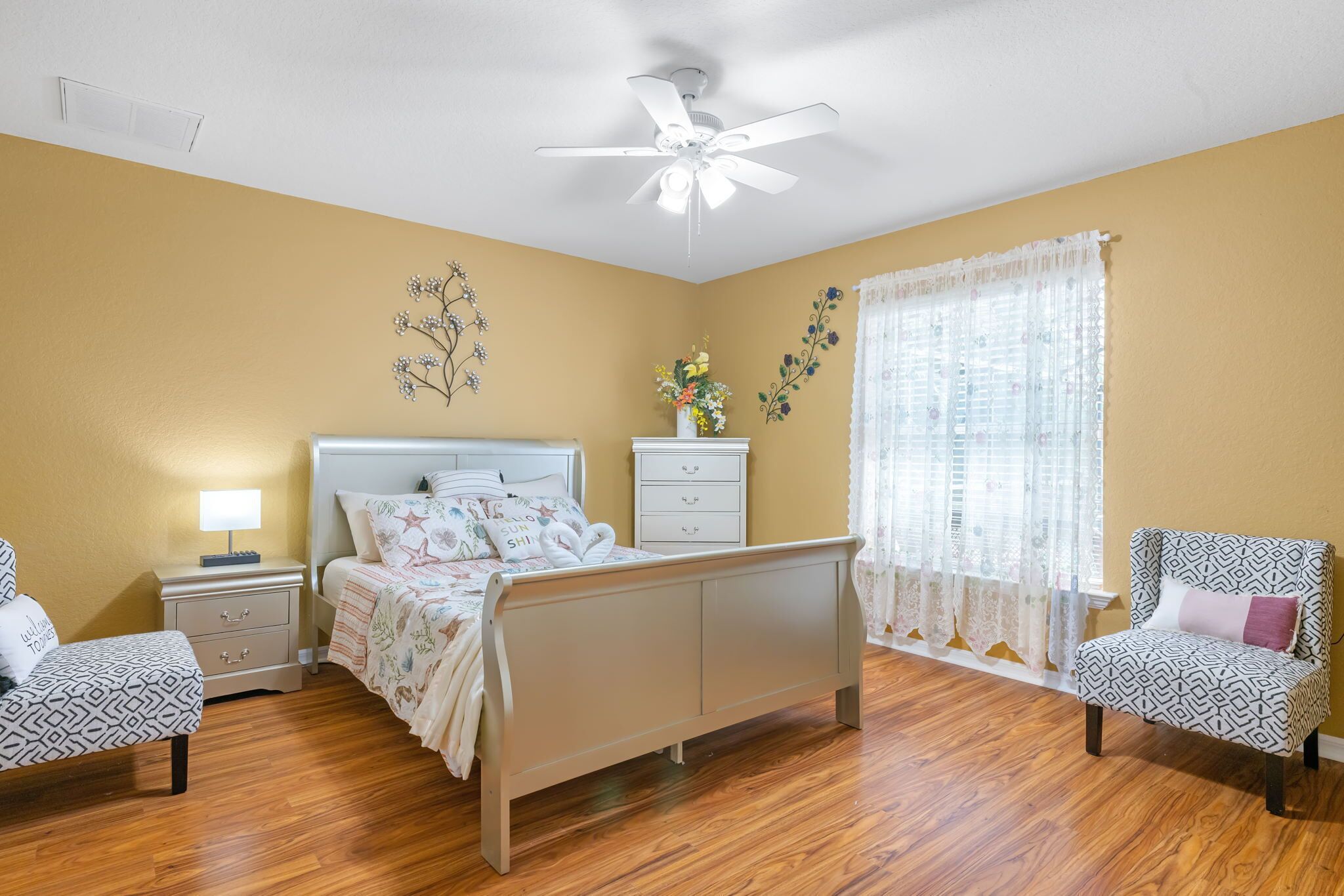 Une chambre à coucher comprenant un lit, des tables de chevet, une chaise, un ventilateur de plafond et une fenêtre avec des rideaux.