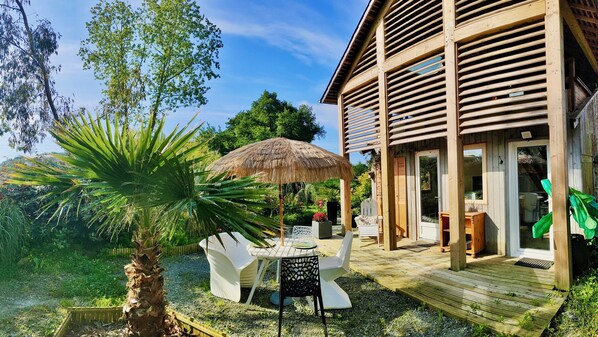 A wooden building with a covered patio area, a table with chairs, and a thatched umbrella.