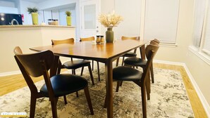 Une salle à manger avec une table en bois et six chaises, un vase contenant des fleurs séchées, et une cuisine en arrière-plan.