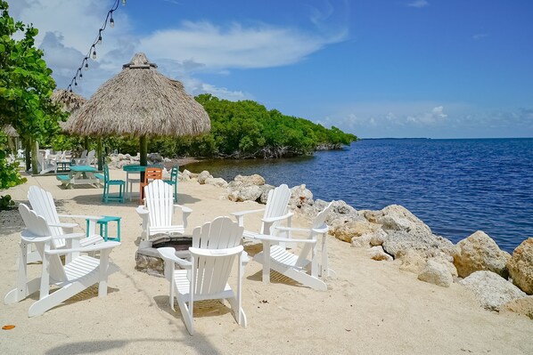 A beachside seating area with white chairs and a thatched umbrella, overlooking a calm sea and a lush green island.