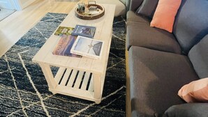 Ein modernes Wohnzimmer mit einem dunkelgrauen Sofa, einem Couchtisch mit Büchern, einem schwarz-weißen Teppich und Blick in den Küchenbereich.