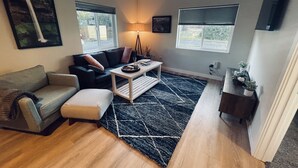 Ein Wohnzimmer mit einer Couch, einem Couchtisch und einem Teppich. Es gibt Fenster, eine Lampe und ein an der Wand befestigtes Fernsehgerät.
