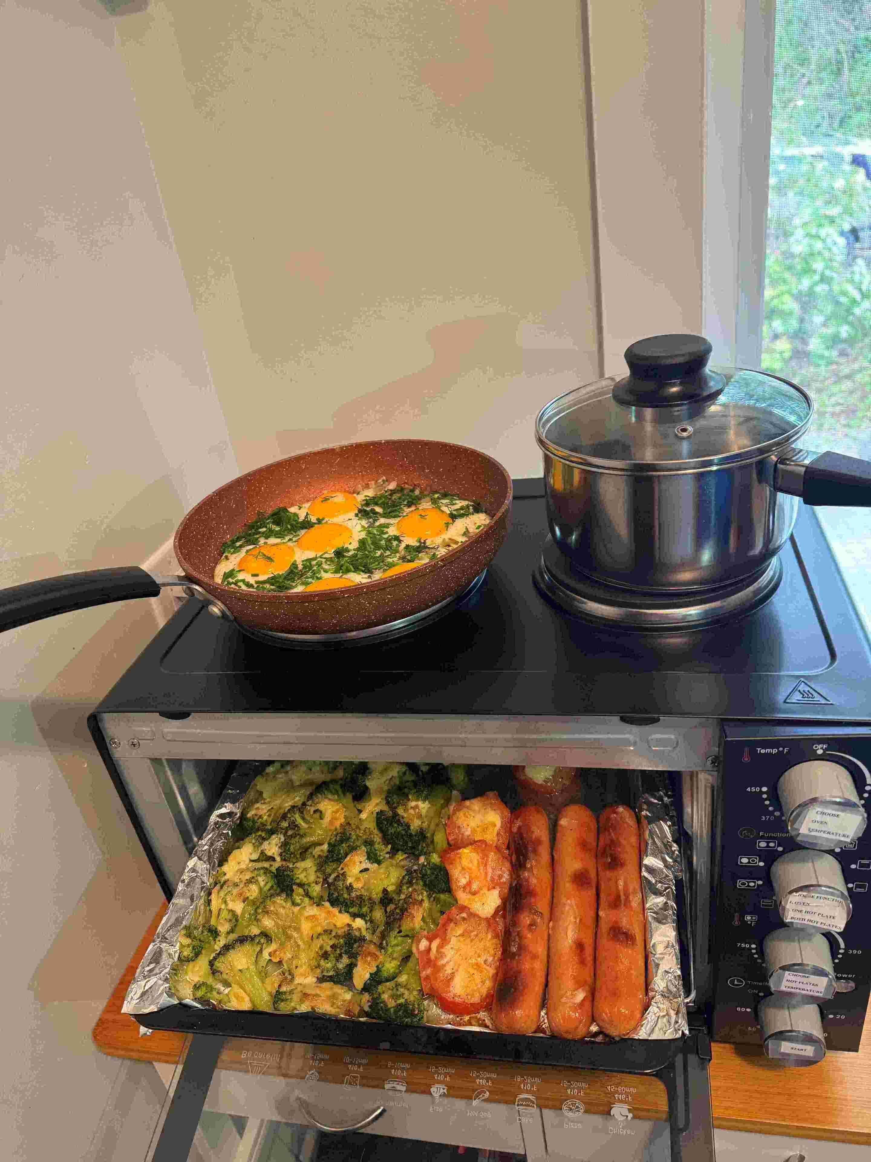 A kitchen with a stove, a pan with eggs, a pot, and a toaster oven containing vegetables and sausages.