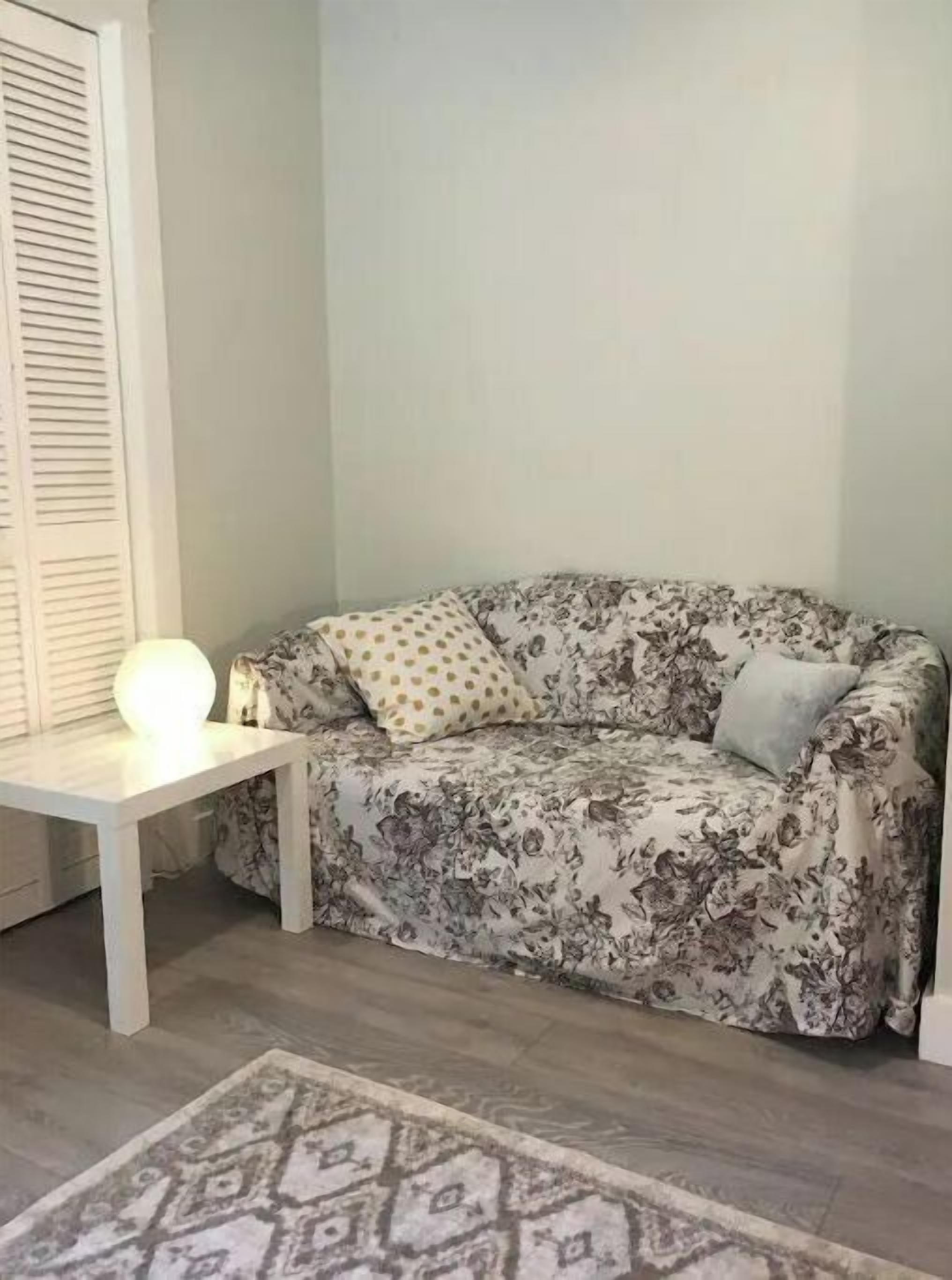 Una habitación con un sofá estampado de flores, una mesita blanca con una lámpara y una alfombra con diseño.