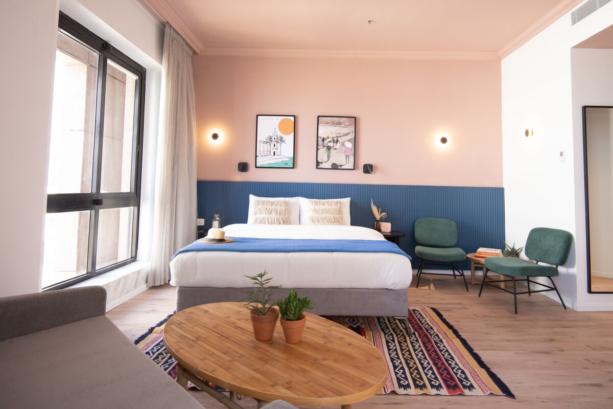 Une chambre d’hôtel moderne avec un grand lit, une table ronde en bois, deux chaises vertes et une grande fenêtre.