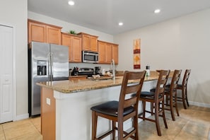 A kitchen with wooden cabinets, a stainless steel refrigerator, a microwave, a sink, and bar stools.