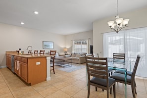 Una cocina moderna con una isla central, un comedor con mesa de vidrio y una sala de estar con sofá y televisor.