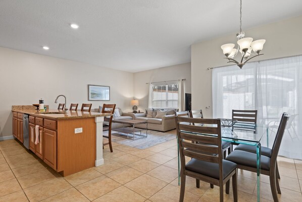 A modern kitchen with a central island, a dining area with glass-top table, and a living room with a sofa and a TV.