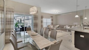 A modern kitchen with a central island, a dining area with a glass table, and a living area with a sofa and curtains.
