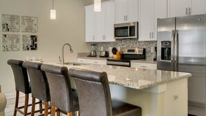 Cocina moderna con armarios blancos, electrodomésticos de acero inoxidable, encimera de granito y una zona de bar con sillas de cuero.