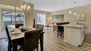 Una cocina moderna con una isla central, comedor con sillas y una mesa dispuesta para una comida, y una sala de estar con sofá y mesa de centro.