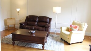 Una sala de estar con un sofá de cuero marrón, un sillón color crema, una mesa de centro de madera y dos lámparas de pie.