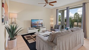 Ein Wohnzimmer mit Sofa, Couchtisch, Fernseher, Deckenventilator und einem großen Fenster mit Vorhängen.