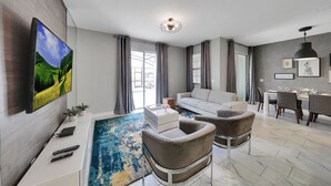 Een moderne woonkamer met een flatscreen televisie, een bank, twee fauteuils, een salontafel en een eethoek met een tafel en stoelen.