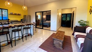 A modern kitchen with bar stools, a dining table, and a large window. There is a living area with a sofa and a coffee table. - Digital Nomad's Dream in Paradise I (Sayulita)