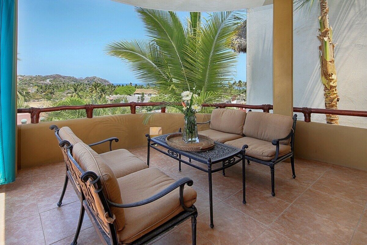 Un balcon avec un canapé et des fauteuils, une table avec un vase, et une vue sur des palmiers et des collines.