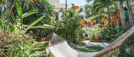 A hammock in a lush garden with tropical plants and a building in the background.