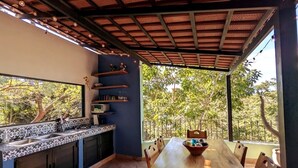 An outdoor kitchen with a dining table and chairs, surrounded by a covered area with a view of trees.