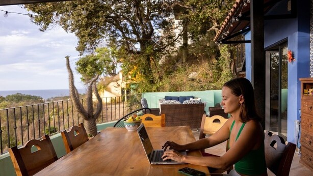 A woman using a laptop on a wooden table in an outdoor setting with a view of trees and a building in the background.