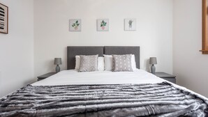 A neatly arranged bedroom with a large bed, two bedside tables, and three framed potted plants on the wall. - West Sands - Donnini Apartments (Ayr)