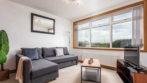 A modern living room with a grey sofa, a small table, a TV, and a large window with a view of the outdoors. - West Sands - Donnini Apartments (Ayr)