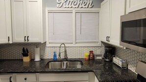 Una cocina con gabinetes blancos, un lavamanos de acero inoxidable, un microondas y una ventana con persianas.