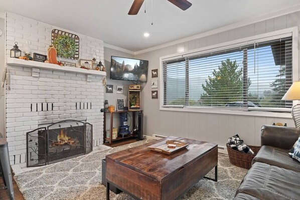A living room with a fireplace, a television, and a large window with blinds. - Rustling Pine Cabin (Greer)