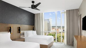Une chambre d’hôtel avec deux lits, un ventilateur de plafond, une grande fenêtre donnant sur un paysage urbain et un téléviseur à écran plat.