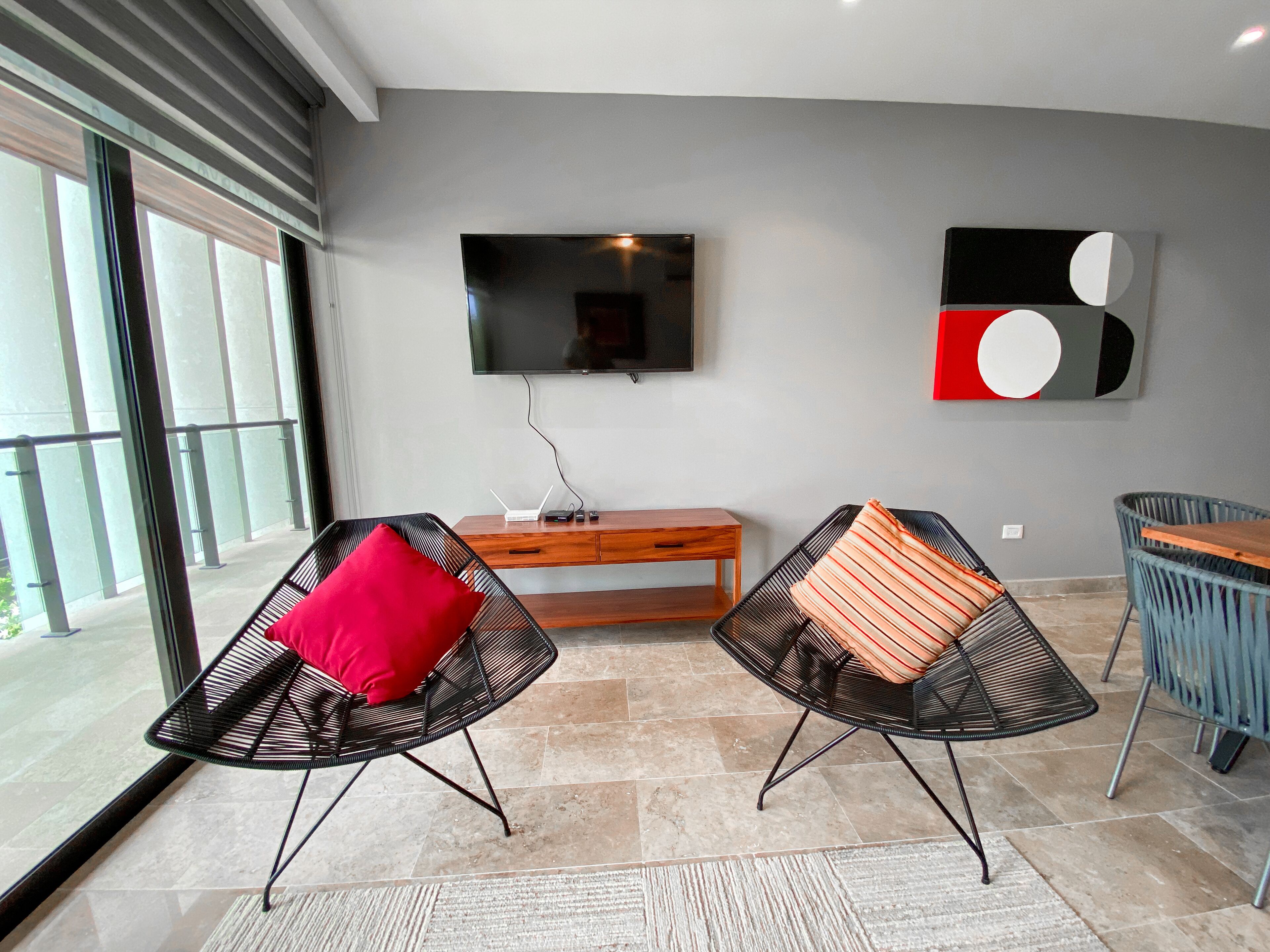 A modern living room with two black wire-framed chairs, a wooden side table, a flat-screen TV, and a large abstract painting.