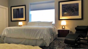 A bedroom with a bed, two bedside lamps, a chair, a window, and two framed pictures on the wall. - Petaluma Warehouse Loft Collection (Petaluma)