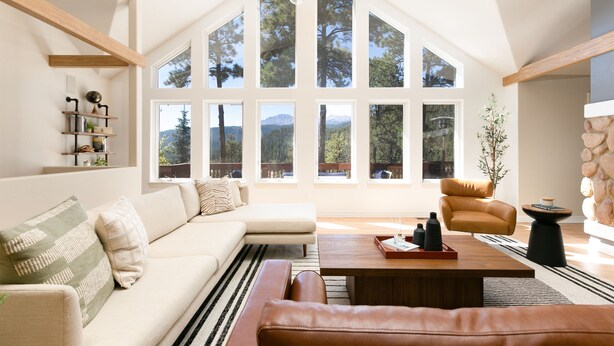 Un salon spacieux avec un plafond haut, de grandes fenêtres offrant une vue sur les montagnes, un canapé en cuir, une table basse et un mur décoratif en pierre.