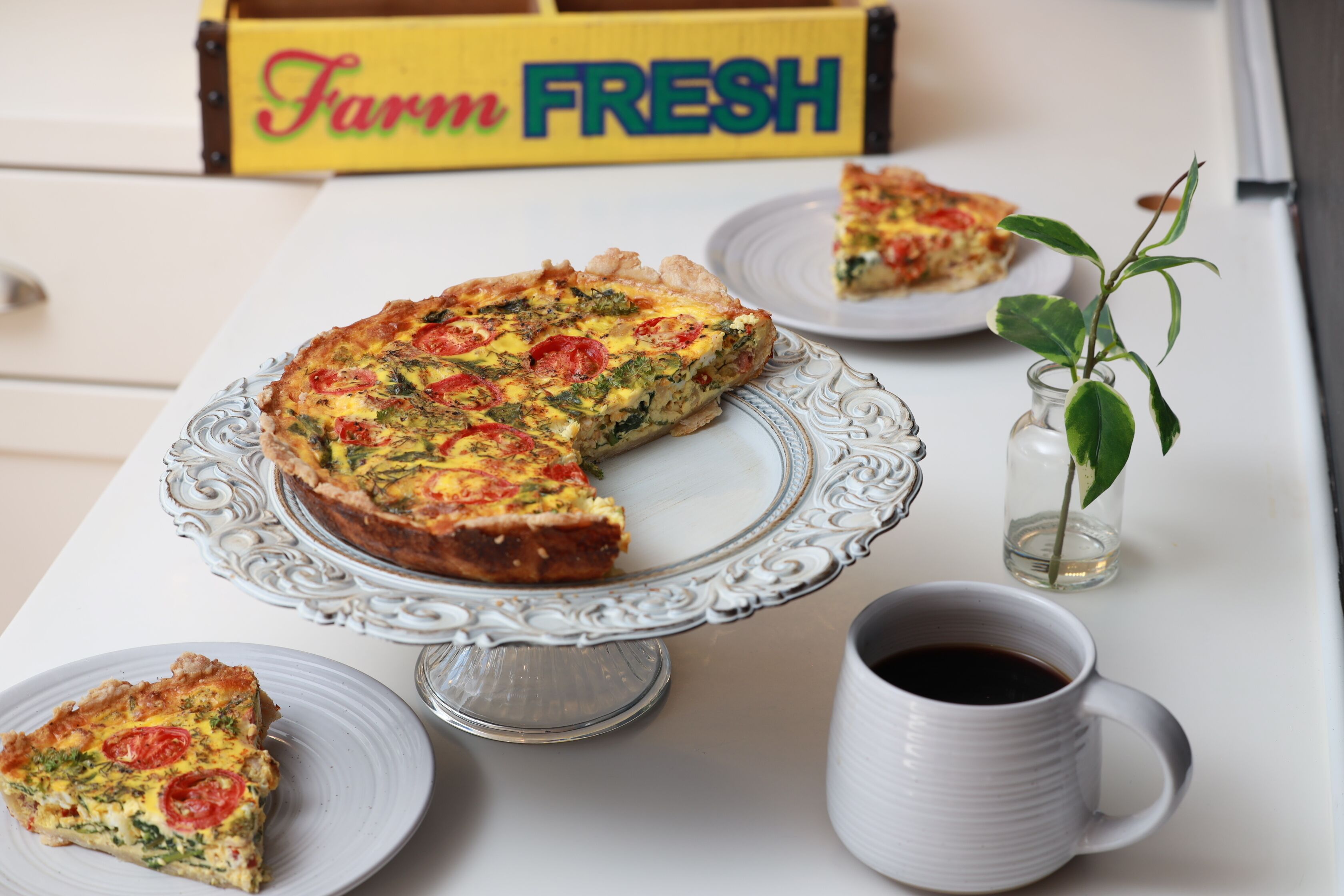 A quiche on a decorative plate, a slice on a smaller plate, and a cup of coffee on a table.