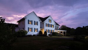 Une grande maison blanche à deux étages, avec un toit foncé, entourée d’une pelouse bien entretenue et d’arbres.