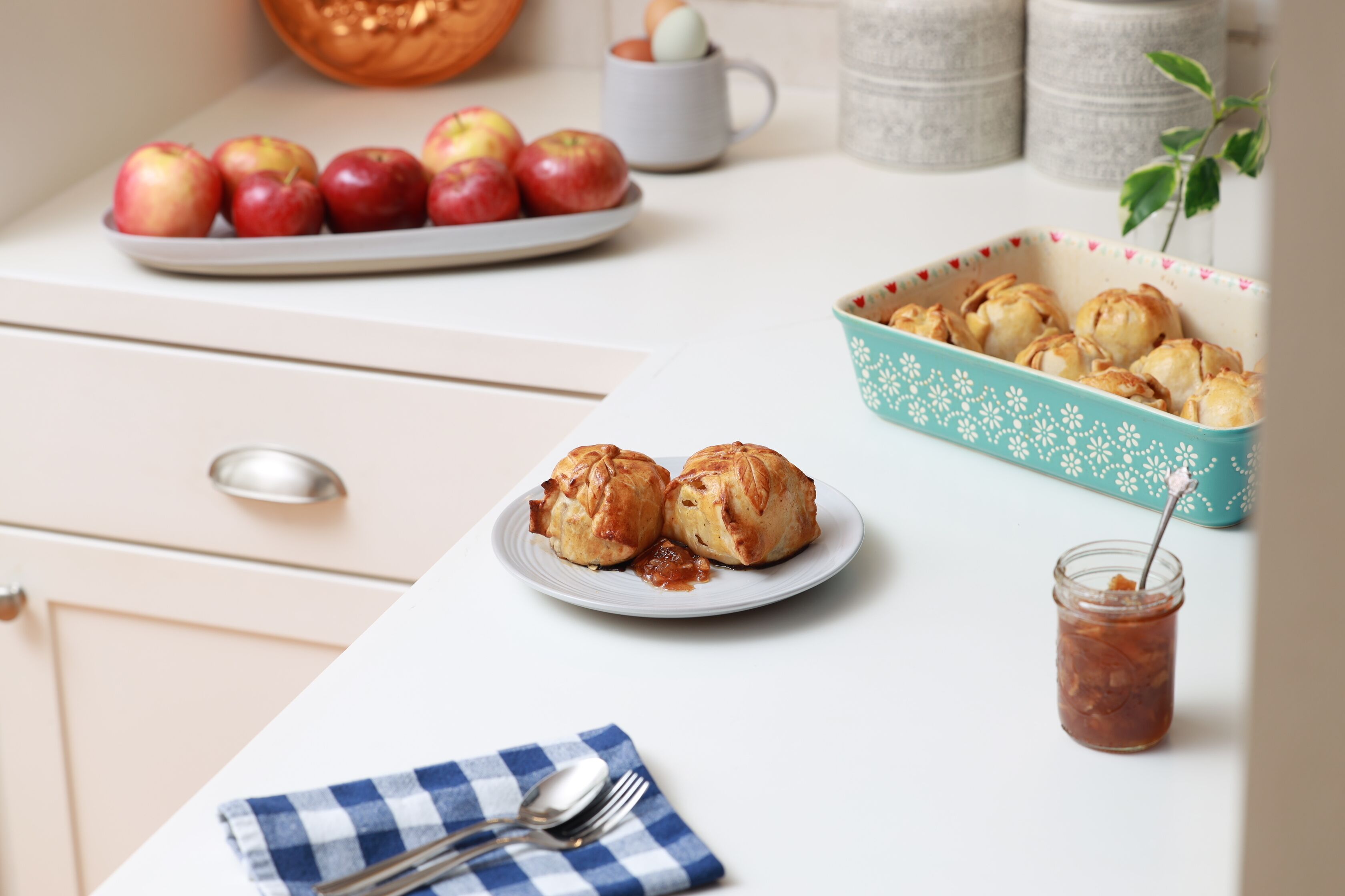 Una encimera de cocina con un plato de manzanas, un bol de pasteles y un frasco de mermelada.