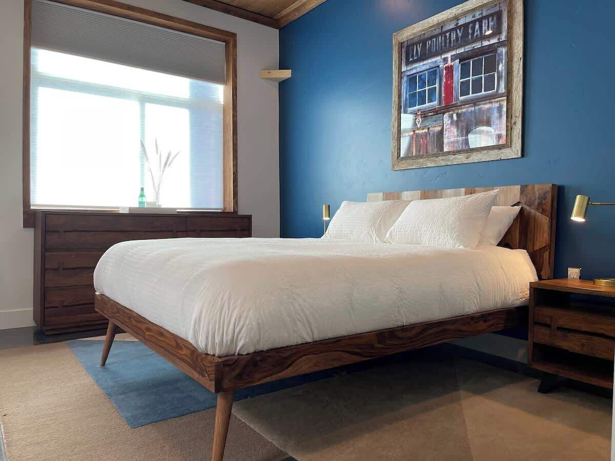 Un dormitorio con cama de madera, mesita de noche, lámpara de pared y un amplio ventanal con persianas.