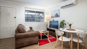 A modern living room with a brown leather sofa, a small wooden table, and a white dining table with chairs. There is a TV on a stand, a lamp, and a potted plant.