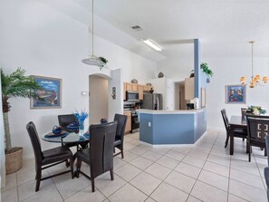 A modern kitchen with a central island, white walls, and a dining area with a glass table and chairs. - Oasis Key Retreat (10 mins from Disney) 75" TV | Private Pool | Game Room (Kissimmee)
