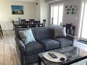 A living room with a grey sofa, two matching cushions, a coffee table with a remote control, and a dining area in the background. - 2BR/2BA LAKEFRONT CONDO 305 Private Beach/Balcony/indoor pool. (Geneva)