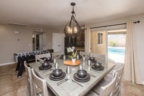 A dining area with a rectangular table set for six, surrounded by chairs. A chandelier hangs above the table, and a foosball table is visible in the background. - “THE SANDY WAKE”Desert Oasis Sleeps 8 with a pool (Lake Havasu City)