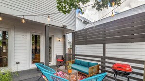 Un patio exterior con terraza de madera, un sofá, dos sillas, una parrilla y luces colgantes.