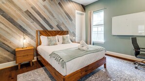 Un dormitorio con cabecera de madera, una mesita de noche, una cama con colcha estampada y un gran ventanal con cortinas.