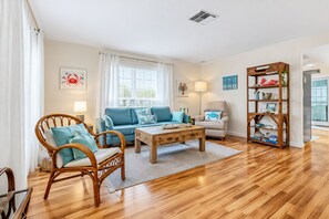 A living room with a wooden floor, a blue sofa, a wooden coffee table, a wicker chair, a white armchair, and a bookshelf.