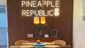 A dining area with a round table, wicker chairs, a wooden wall with vertical slats, and a neon sign reading 'PINEAPPLE REPUBLIC'. - Vibrant Tropical Escape – Just Minutes from the Beach + Serene, Fenced Yard! (Largo)