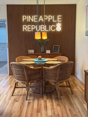 A dining area with a round table, wicker chairs, a wooden wall with vertical slats, and a neon sign reading 'PINEAPPLE REPUBLIC'.