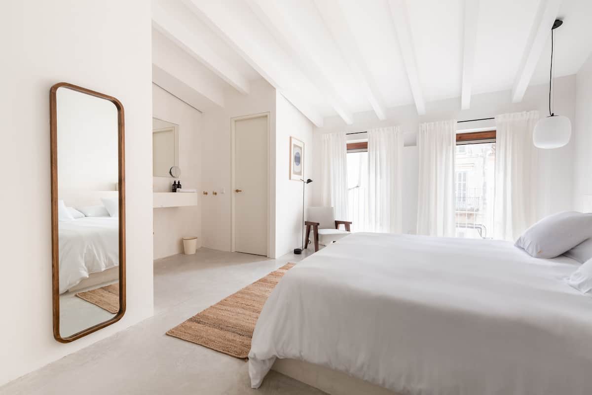 A modern bedroom with a large bed, a mirror, a bedside table, a lamp, and a view of the city through the window.