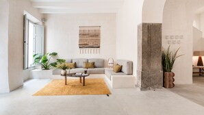 A modern living room with a grey sofa, a coffee table, and a yellow rug. There is a large window on the left and a stone pillar on the right. - Meem Townhouse (Sóller)