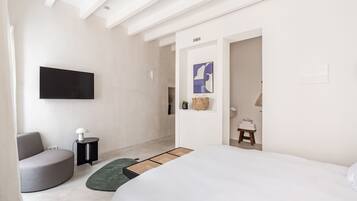 A modern bedroom with a bed, a grey armchair, a small side table, a flat-screen TV mounted on the wall, and a doorway leading to another room.