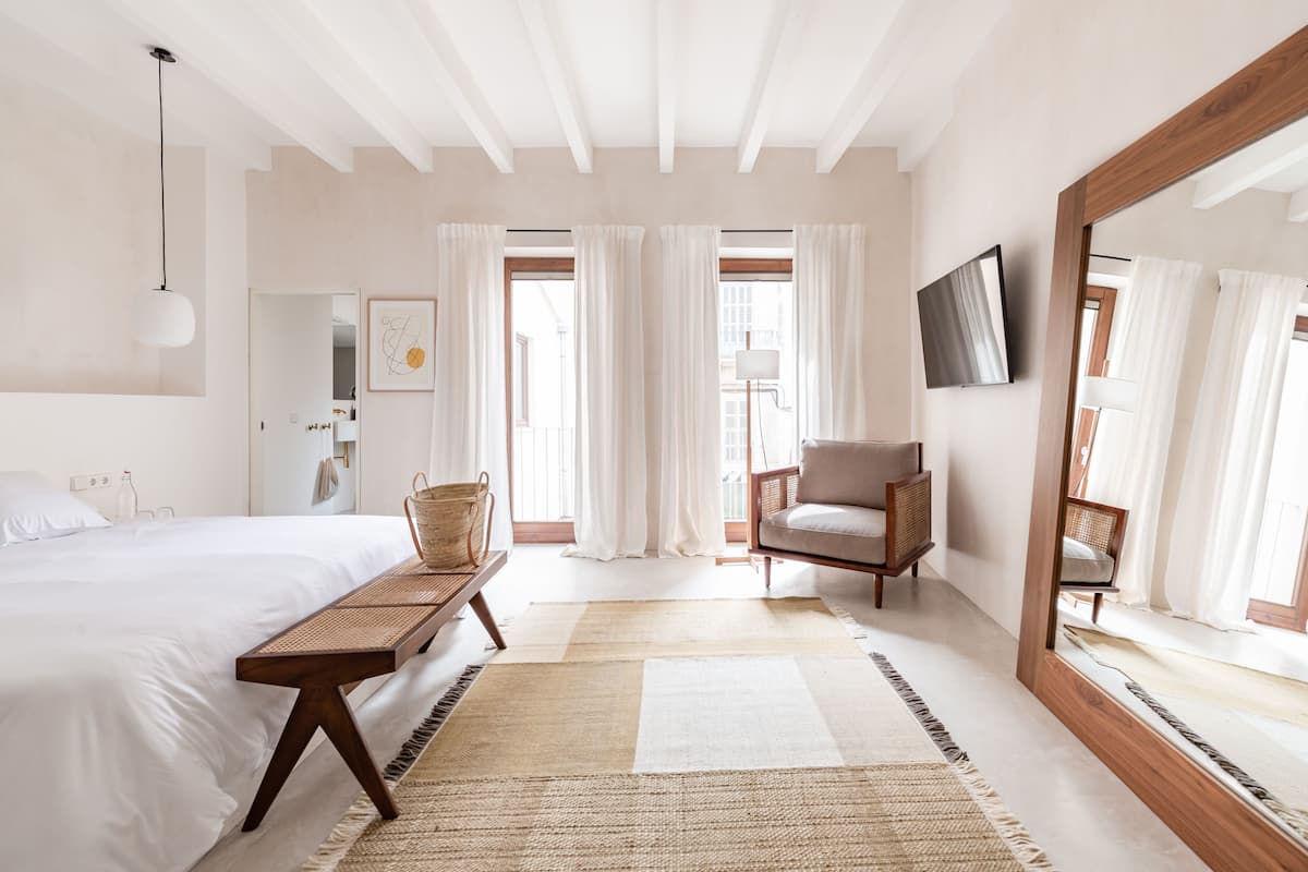 A bedroom with a large bed, a wooden bench, a chair, a TV mounted on the wall, and a large mirror.
