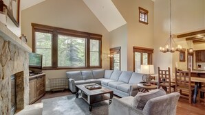 A spacious living area with a stone fireplace, large windows, a grey sectional sofa, and a dining area with wooden furniture. - Elegant 3 Bedroom Townhome with Fireplace (Breckenridge)