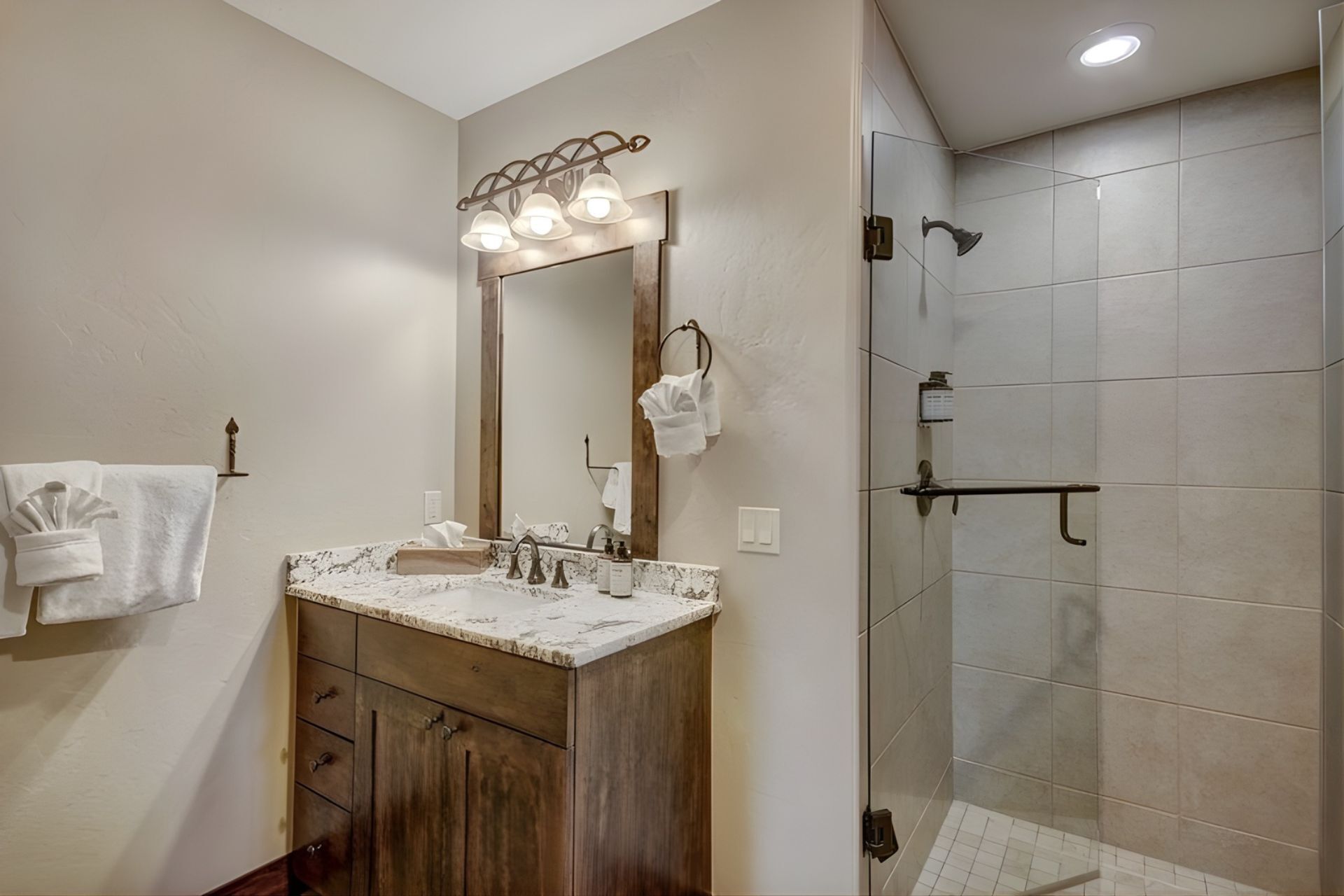 A bathroom with a marble sink, wooden vanity, a large mirror with lights, a towel rack, and a walk-in shower with tiled walls.